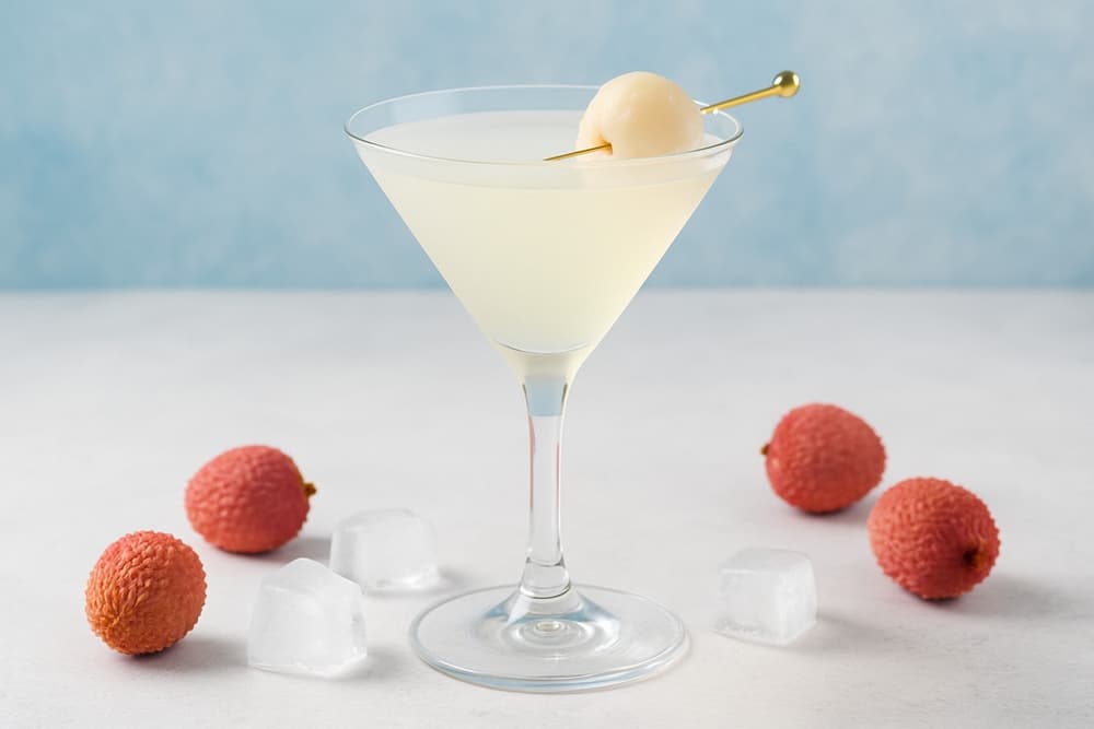 A chilled lychee martini in a clear glass, garnished with a whole lychee, with ice cubes and fresh lychees on a white surface against a soft blue background.