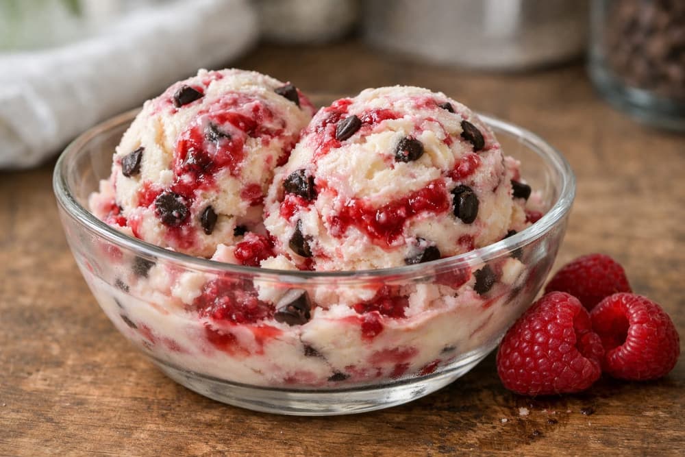 Two scoops of white chocolate ice cream with raspberry swirls and chocolate chips in a glass bowl, raspberries beside