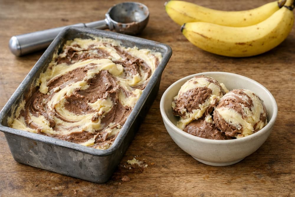 A loaf pan of banana and chocolate ice cream swirled together, with two scoops in a bowl and ripe bananas behind