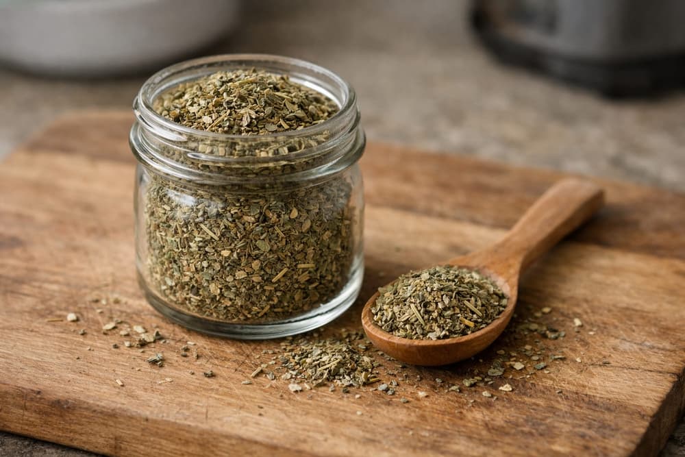 A small glass jar of grey-green dried herb blend on a wooden board, a wooden spoonful of it beside the jar