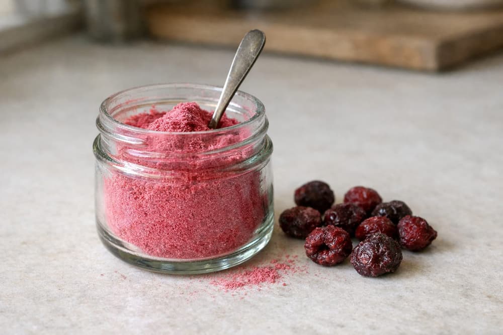 A small glass jar of fine pink cherry powder with a spoon in it, freeze-dried cherries beside the jar